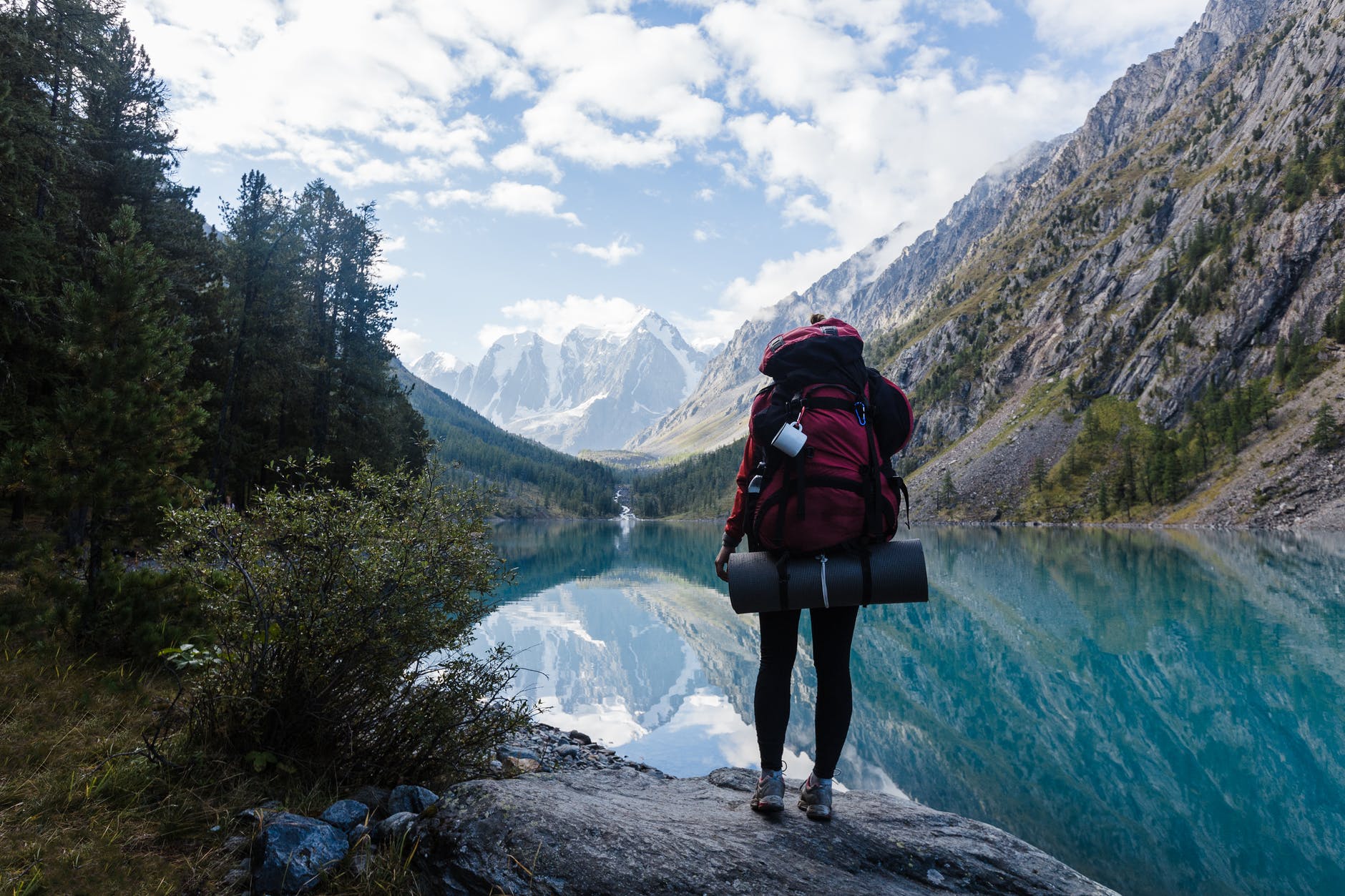 person in red jacket and black pants standing on rock near lake