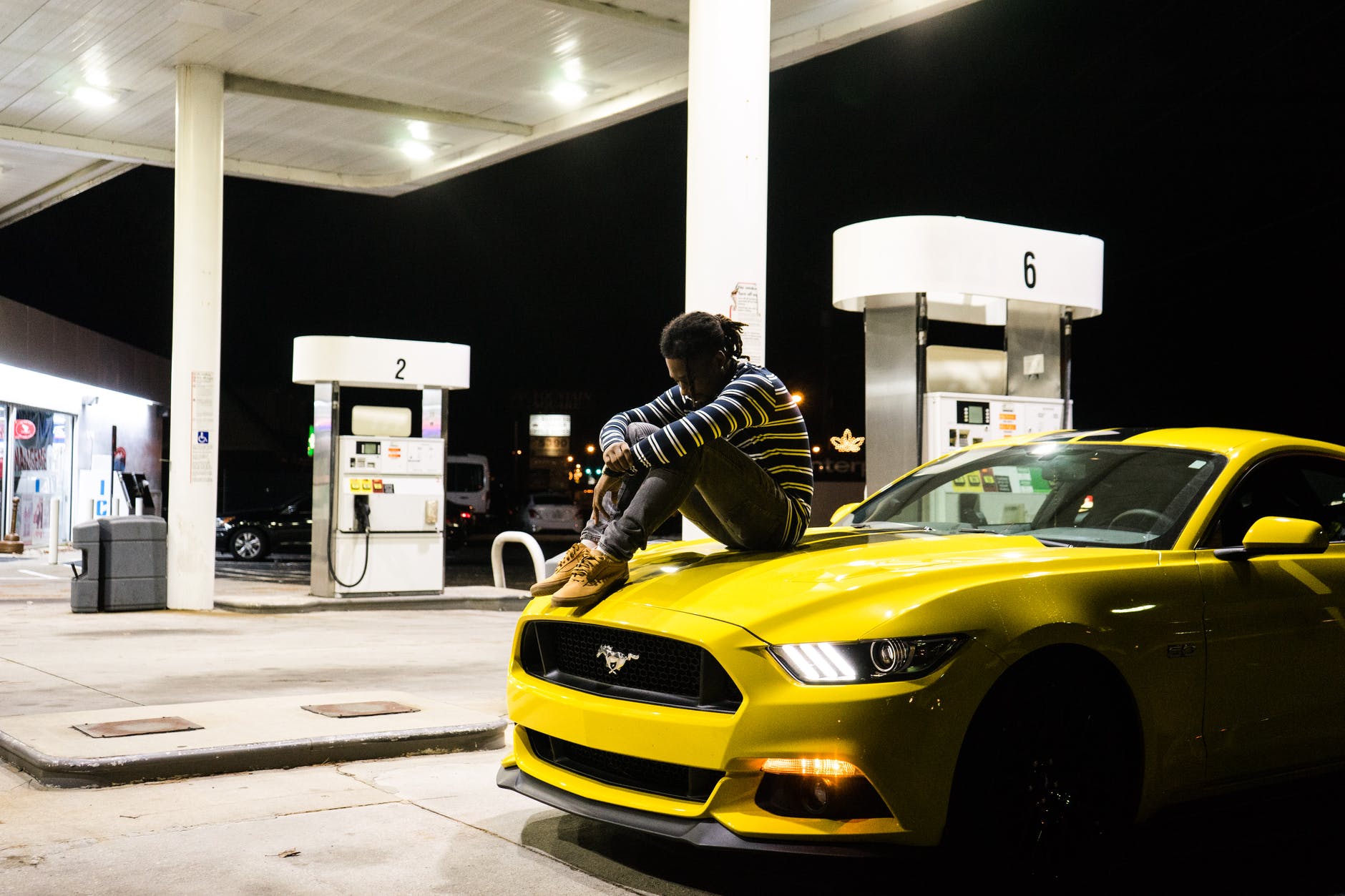 photo of man sitting on hood of yellow ford mustang parked at a gas station